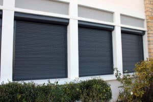 Trois volets roulants noirs fermés sur des fenêtres encadrées de blanc, devant un mur clair et des buissons verts.