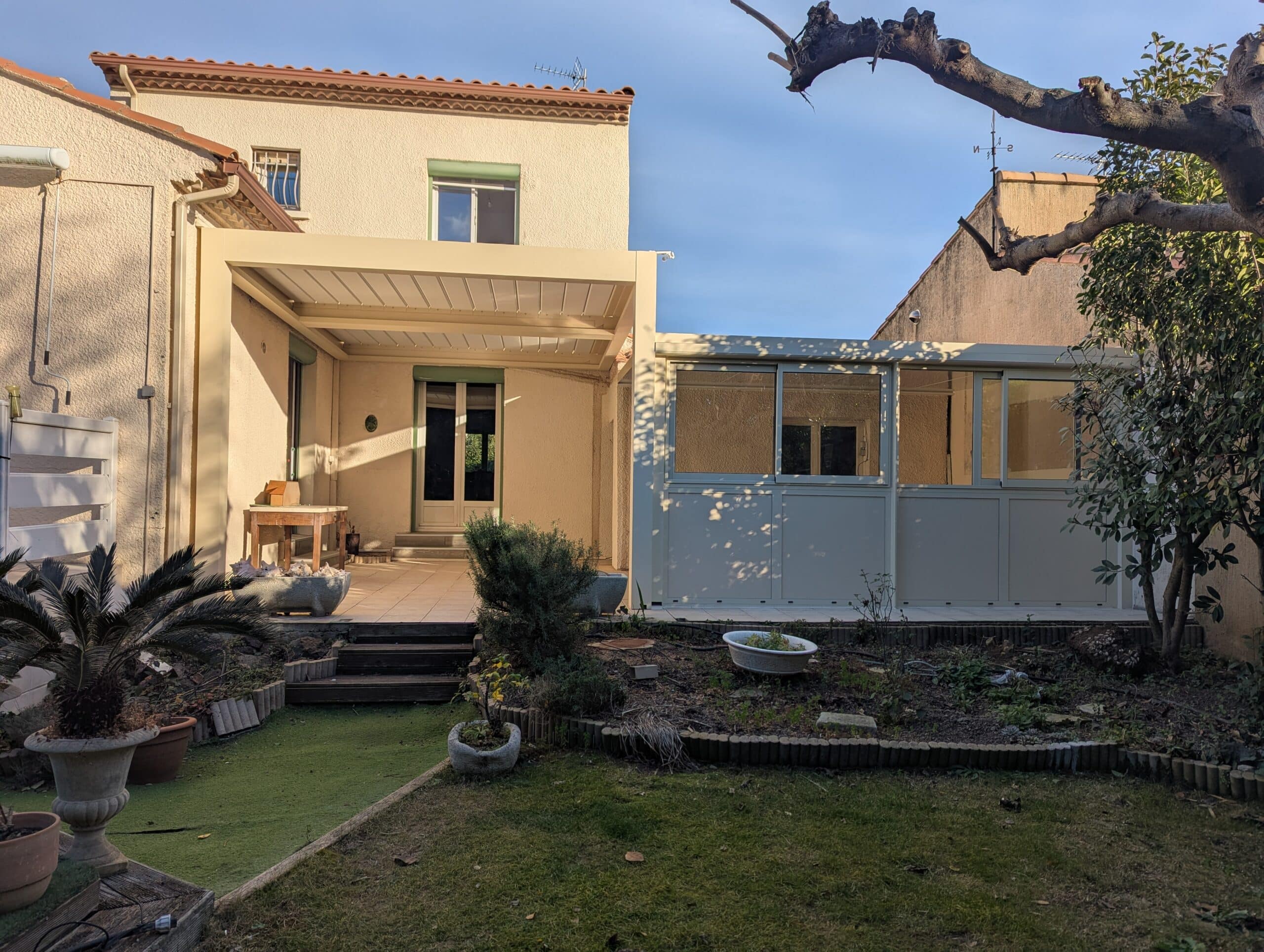Arrière-cour ensoleillée avec patio couvert, véranda moderne et jardin en friche, dominée par les branches d'un arbre taillé.