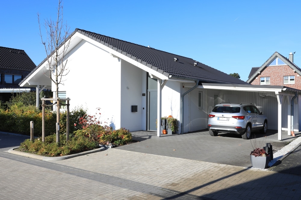 Maison contemporaine blanche avec carport et SUV argenté Maison blanche contemporaine avec toit sombre, carport et SUV argenté stationné sous un ciel bleu.